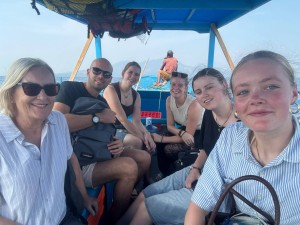 Zes personen poseren onder de overkapping van een blauwe boot, met de zee op de achtergrond.