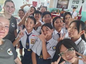 Leerlingen in schooluniform poseren met twee bezoekers in Amurang-shirts voor een groepsselfie in het klaslokaal.
